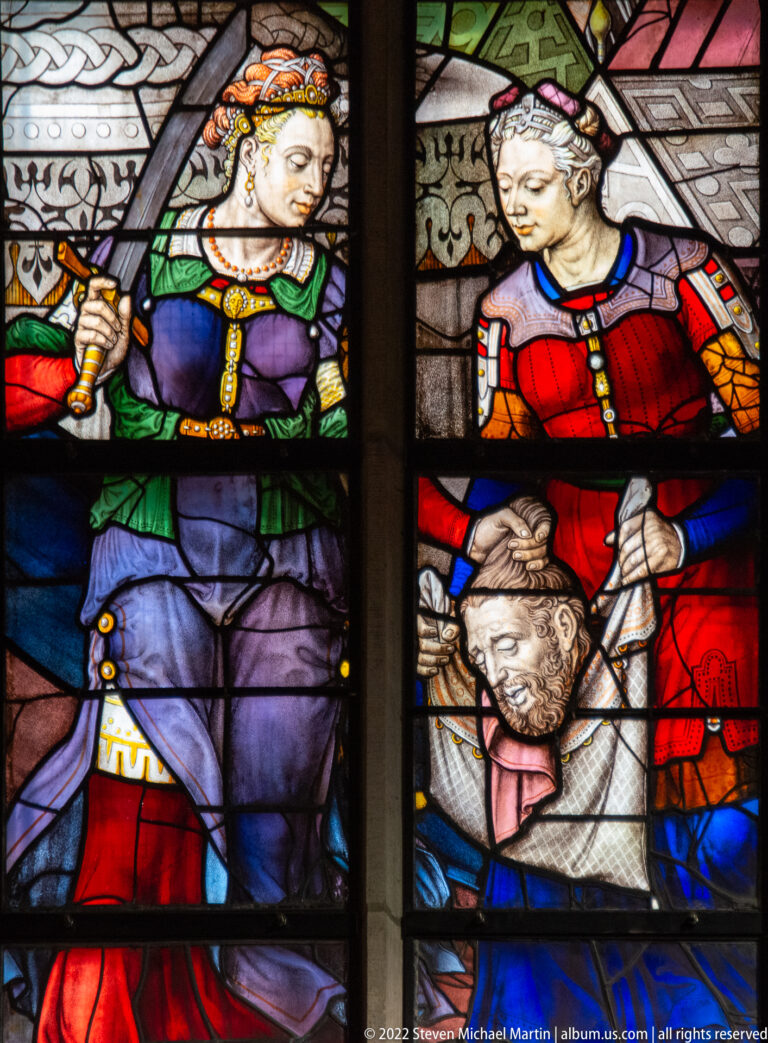 Close stained-glass detail of three richly dressed figures surrounding a bowed bearded man.