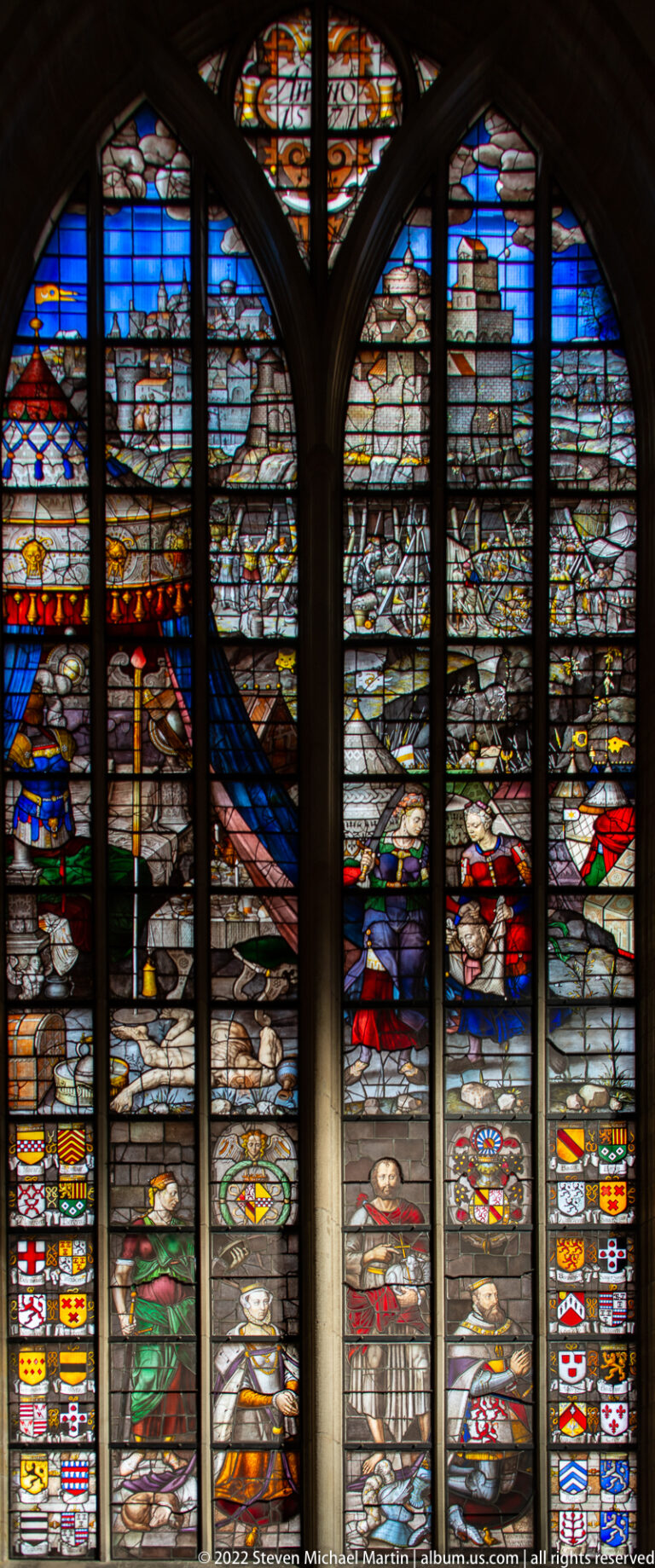 Large arched stained-glass window with several narrative scenes and rows of heraldic shields.