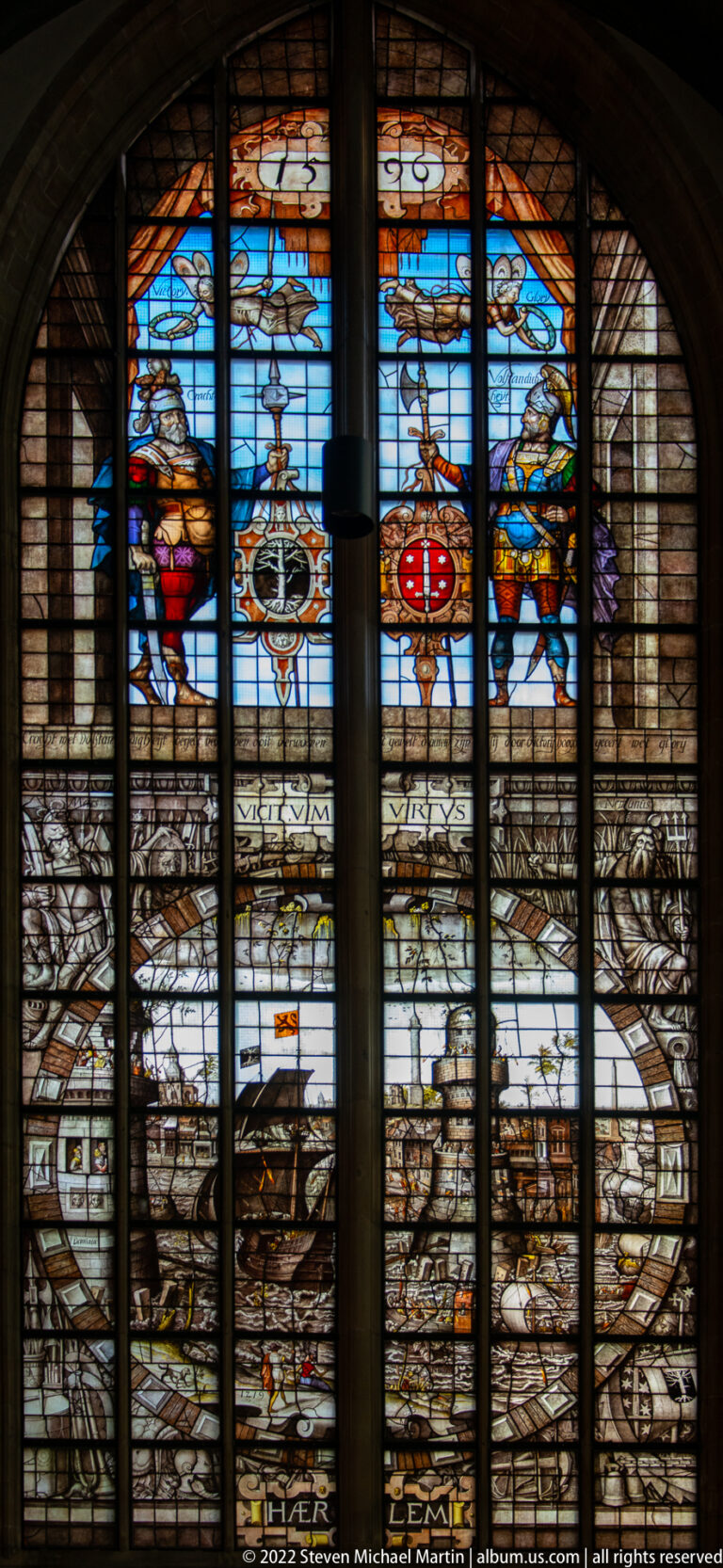 Arched stained-glass window with a pale cityscape below colorful heraldic shields and figures.