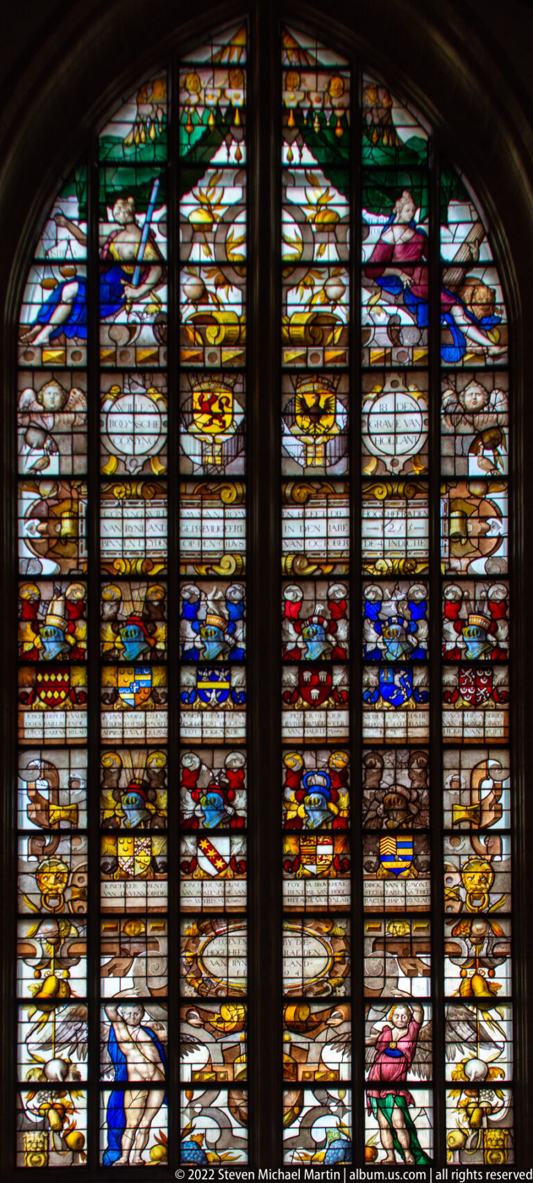 Vertical stained-glass window filled with colorful figures, inscriptions, heraldic shields, and decorative panels.