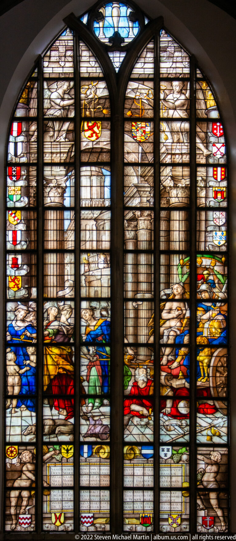 Large stained-glass window with classical columns, arches, figures, and heraldic shields.