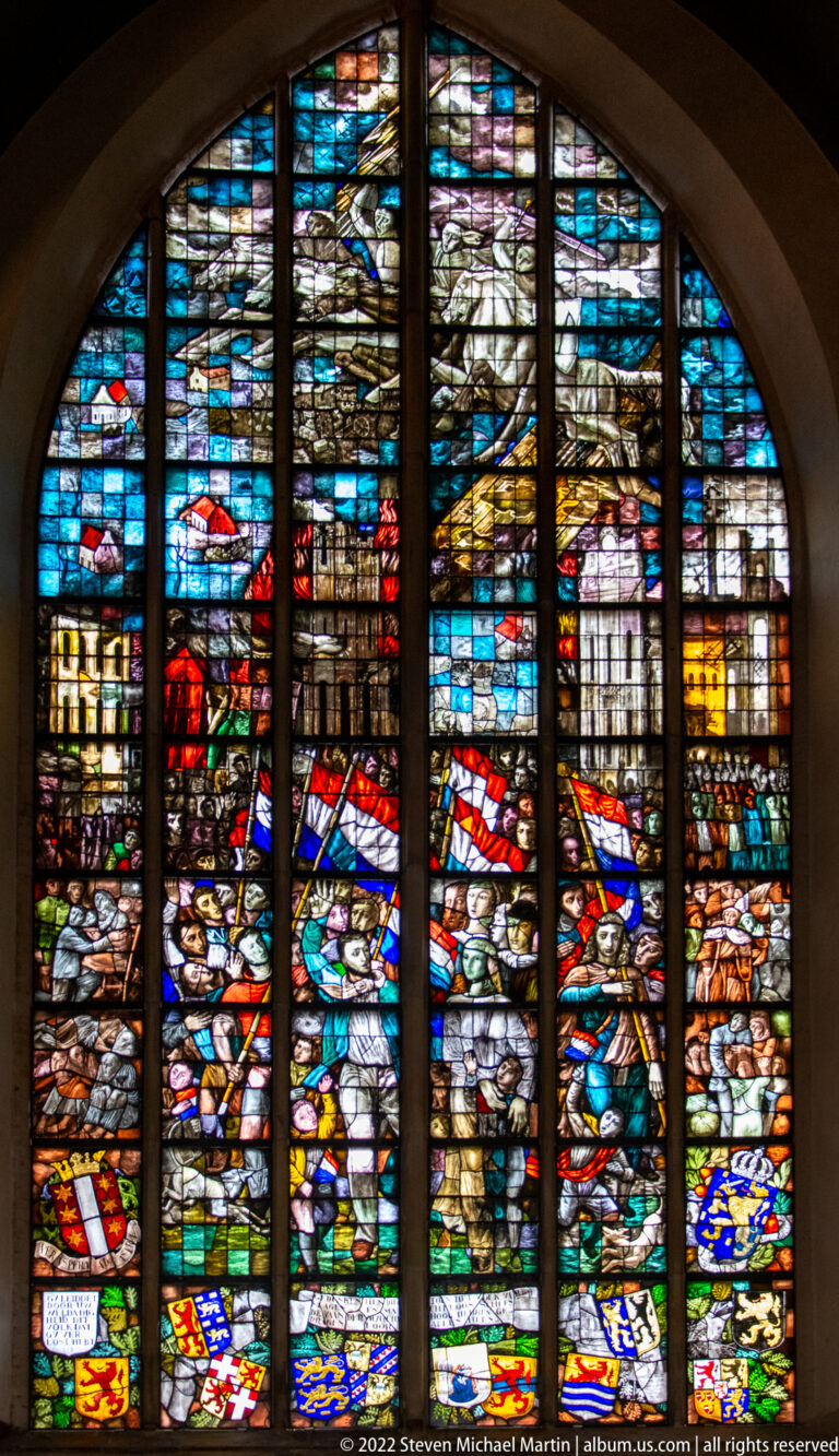 Colorful stained-glass scene crowded with figures, banners, shields, and a bright blue upper background.
