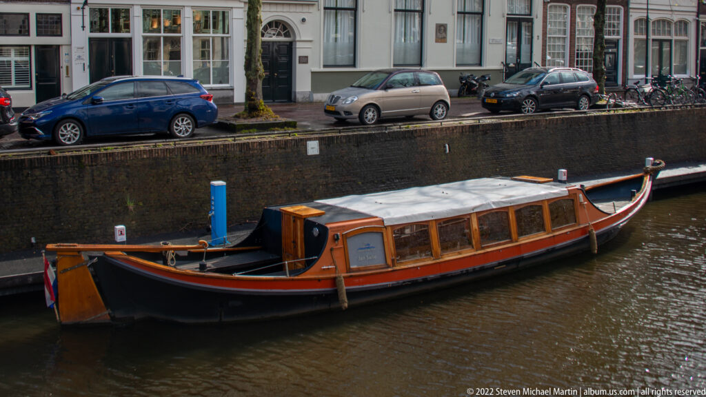 Long covered wooden passenger boat moored on a Gouda canal below parked cars and bicycles.