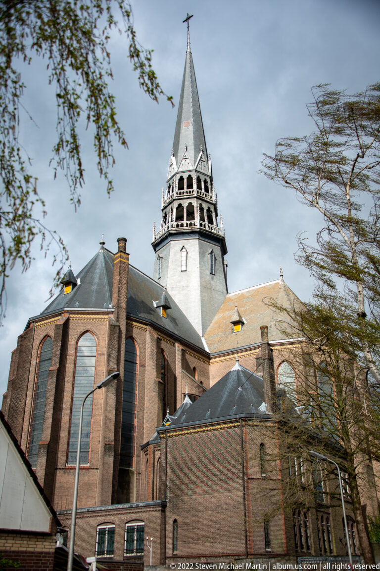 Side exterior of Sint-Janskerk with brick walls, tall pointed windows, roofs, and pale tower.