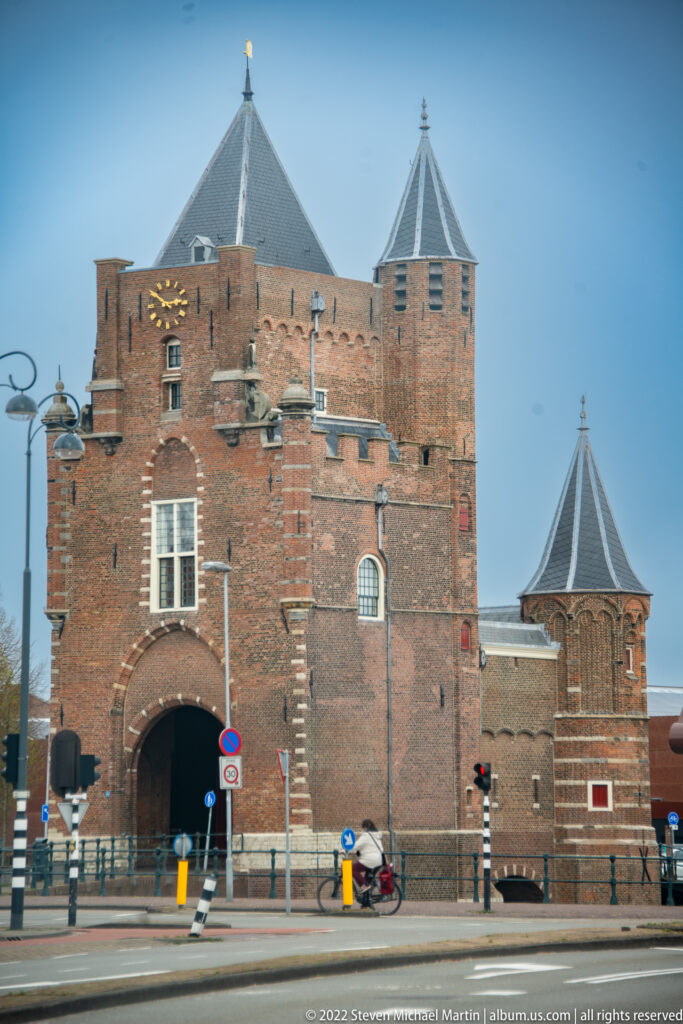 Red-brick Amsterdamse Poort with corner towers, slate roofs, clock, and central passage beside a modern Haarlem street.