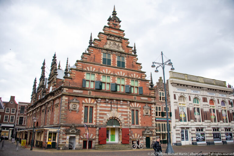 Full street view of the Vleeshal on Grote Markt, with stepped gable, red brick, pale stone bands, bicycles, and neighboring buildings.
