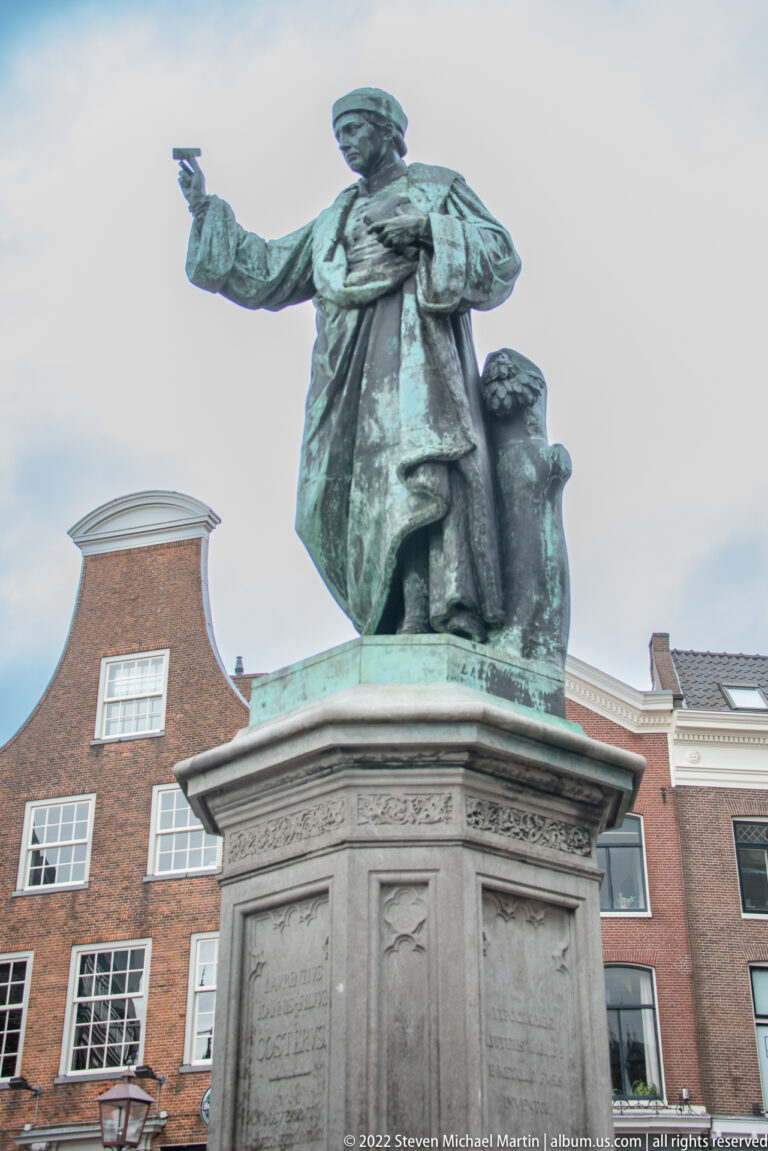 Bronze statue of Laurens Janszoon Coster in a cloak, one arm extended, on a tall stone pedestal in Haarlem.