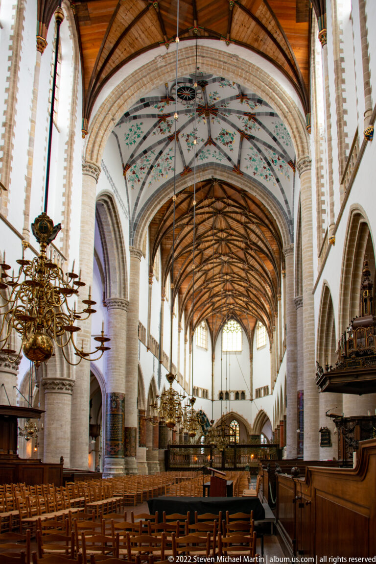 Wide view down the Grote Kerk nave with white arcades, timber vault, brass chandeliers, and rows of chairs.