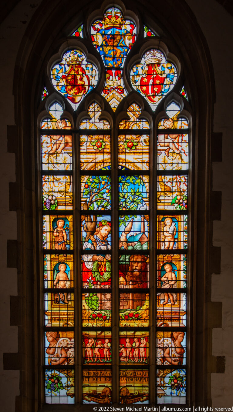 Colorful stained-glass window in a Gothic stone opening, with heraldic shields, figures, and decorative borders.