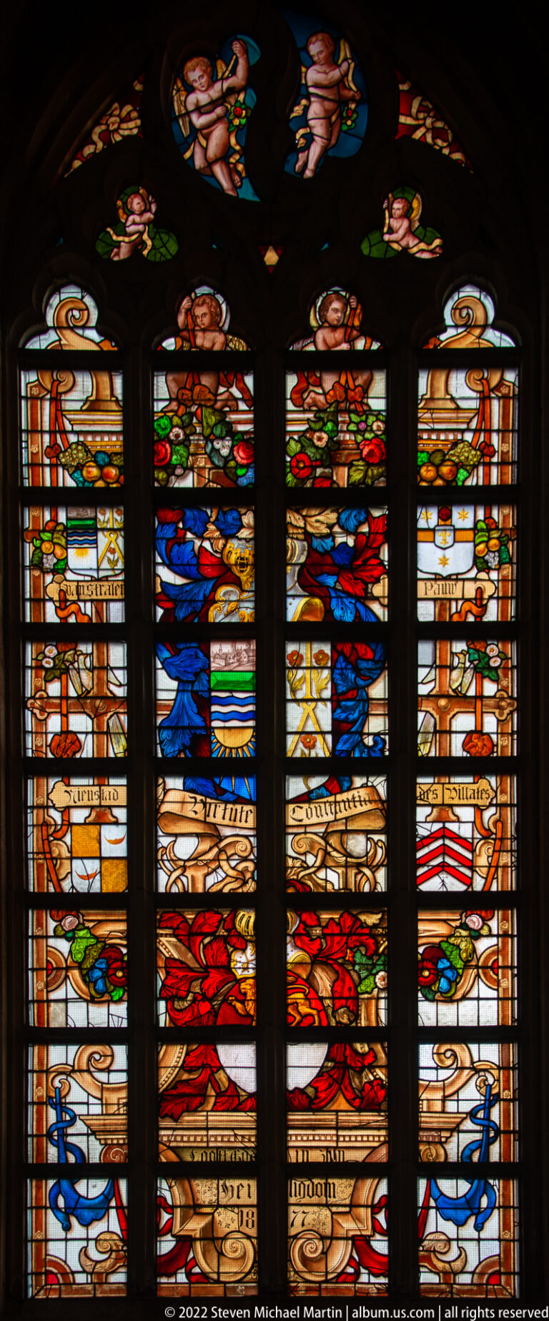 Close view of a stained-glass window with large heraldic shields, smaller figures, inscriptions, and decorative borders.