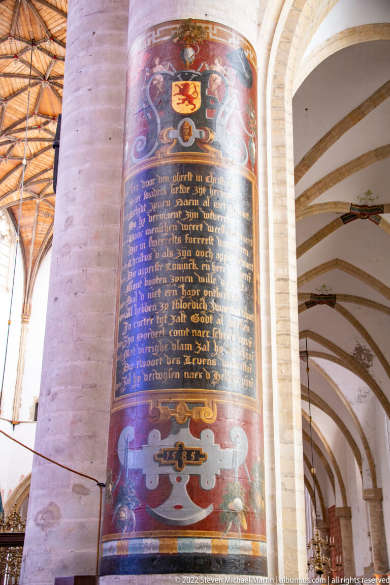 Painted memorial panel on a church pier with Dutch inscription, coats of arms, and decorative framing.