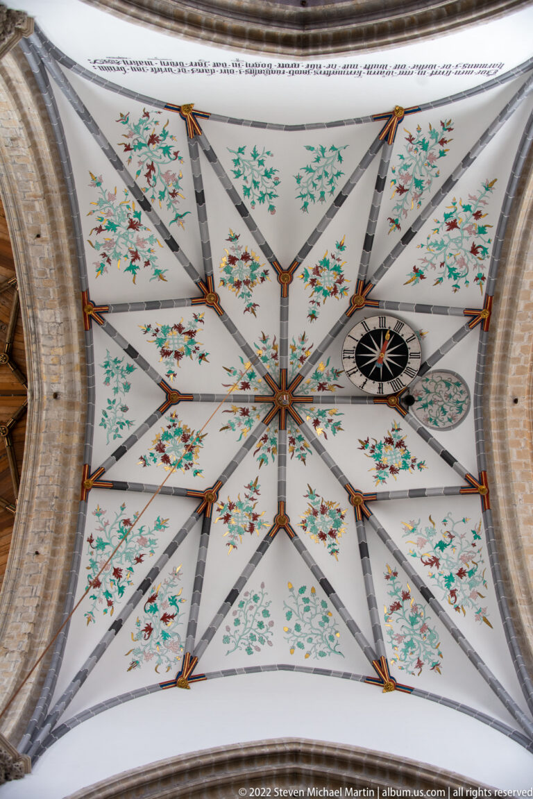 Upward view of a pale church vault painted with green floral motifs, red accents, geometric ribs, and a dark circular dial.