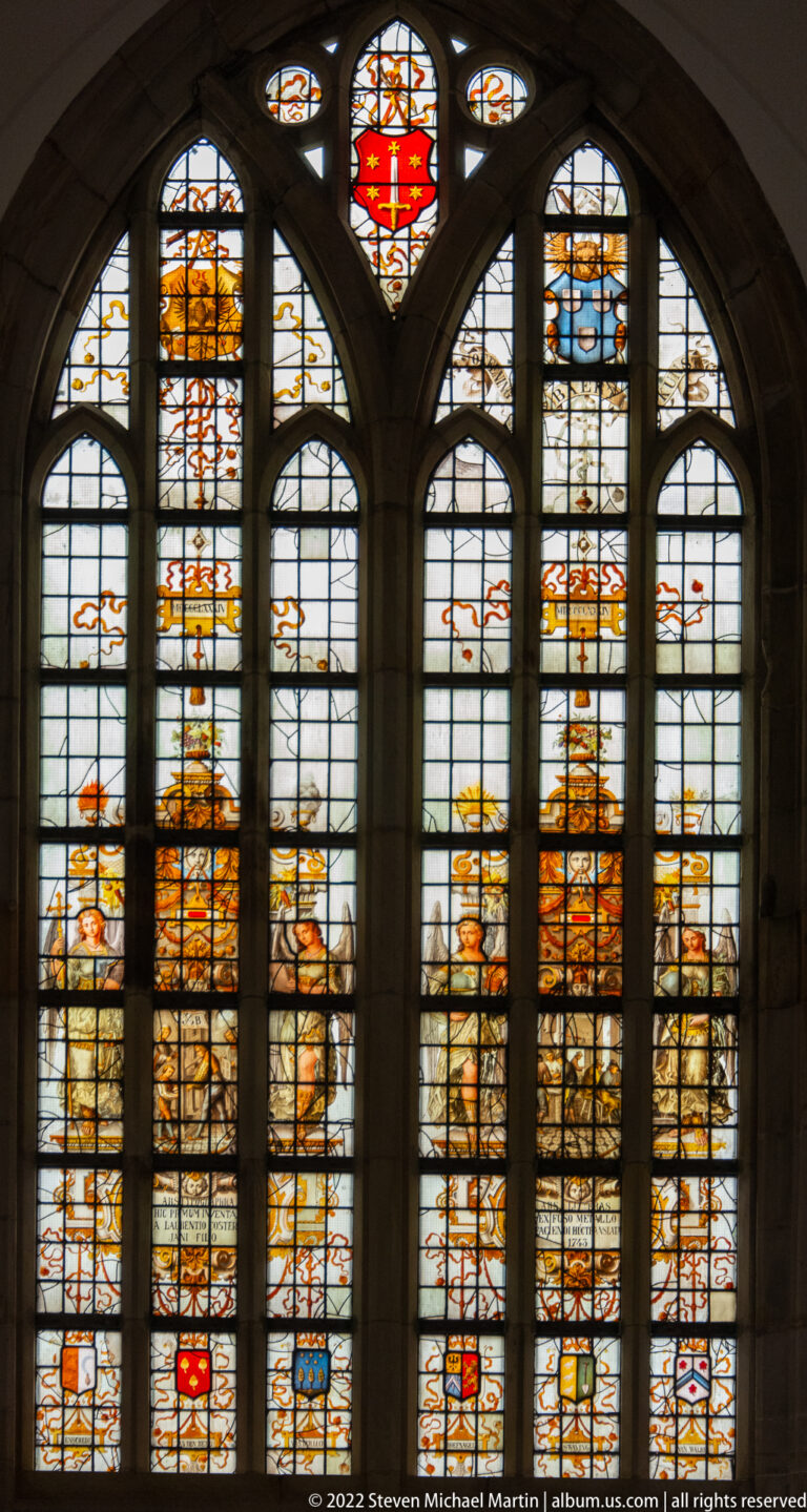 Three tall Gothic windows filled with heraldic stained glass, coats of arms, figures, and amber-colored decorative panels.