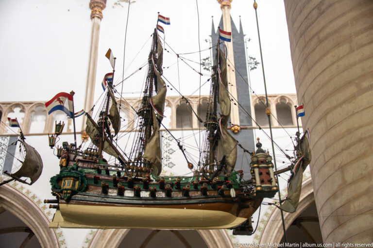 Decorated model sailing ship with Dutch flags and detailed rigging suspended beneath pale church arches.