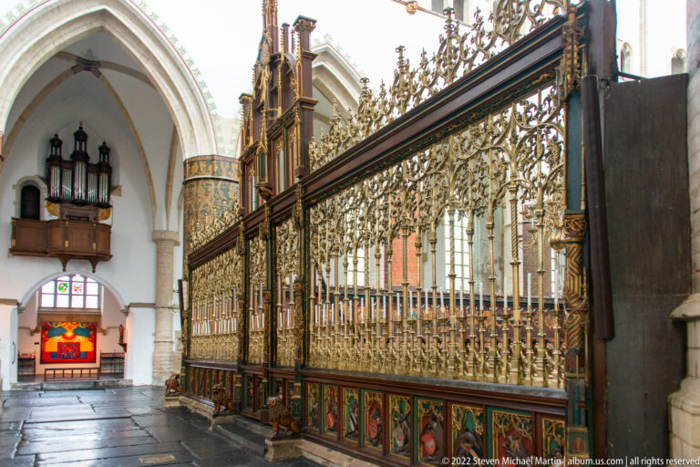 Long ornate metal choir screen with gilded openwork and painted lower panels beside stone columns and a small organ.