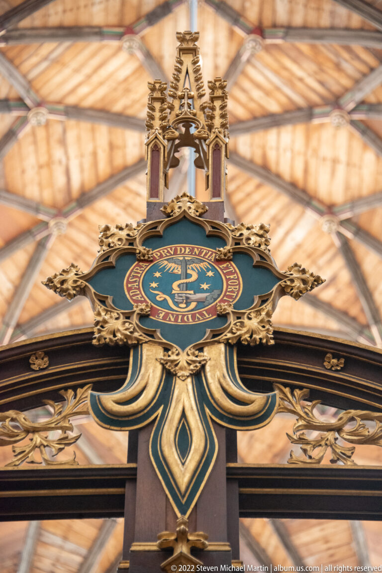 Blue and gold heraldic crest with carved finials mounted above an ornate church screen beneath a wooden roof.