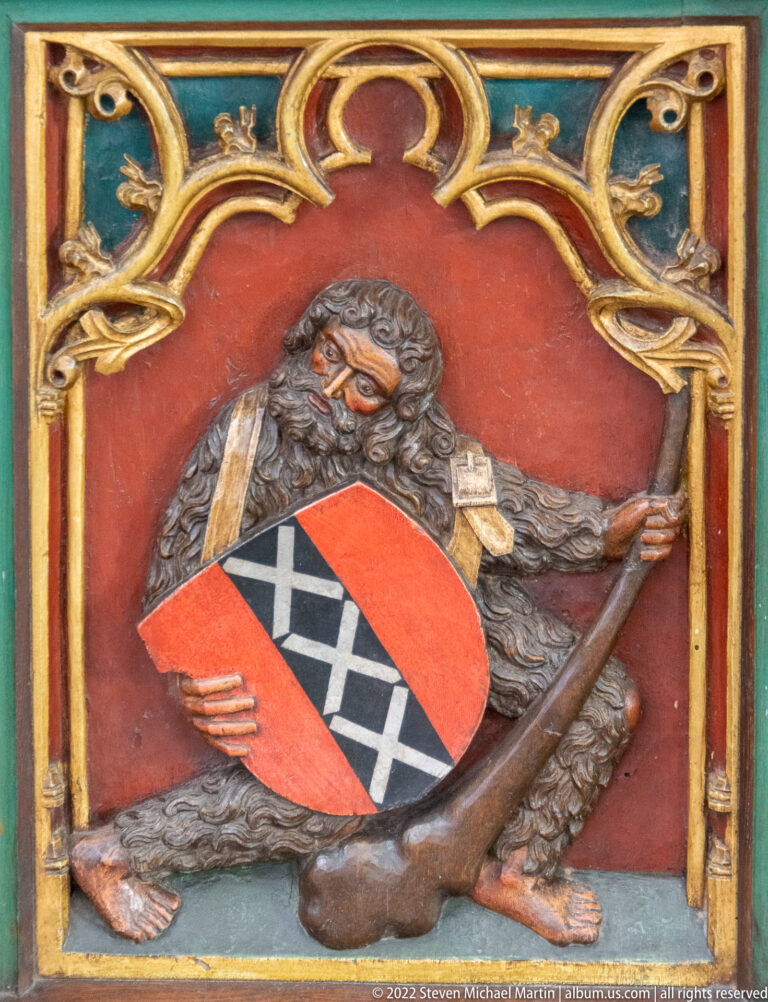 Bearded carved figure in a red niche holding a club and a red, black, and white heraldic shield.