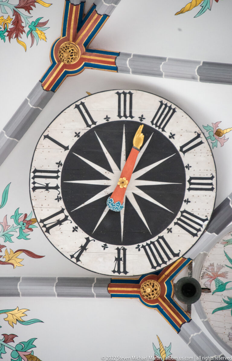 Large decorative clock face with Roman numerals, black center, colored hands, and painted floral ornament around it.