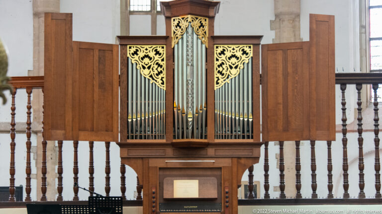Small wooden pipe organ with silver pipes and gilded pierced panels, set against a timber balustrade.