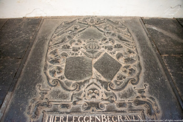 Weathered stone floor grave slab with two shields, scrolling ornament, and an inscription along the lower edge.