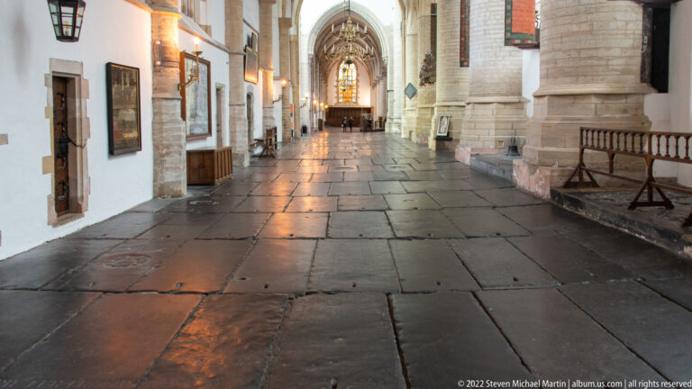 Long stone-paved church aisle lined with massive columns, wall memorials, and a stained-glass window at the far end.