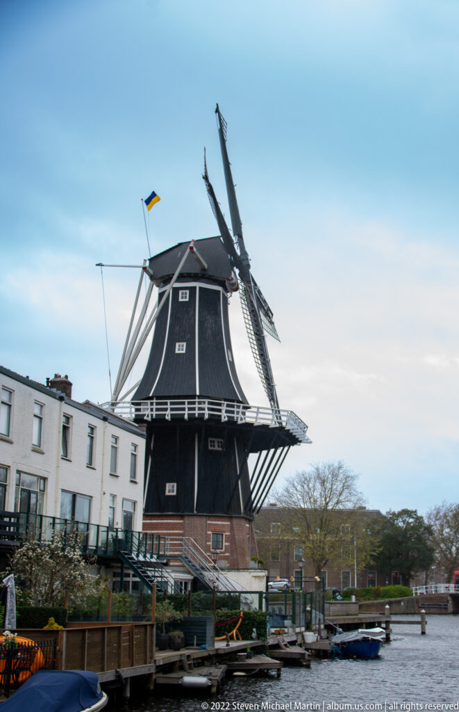 Dark timber windmill De Adriaan with four sails beside a Haarlem canal, low houses, and moored boats.