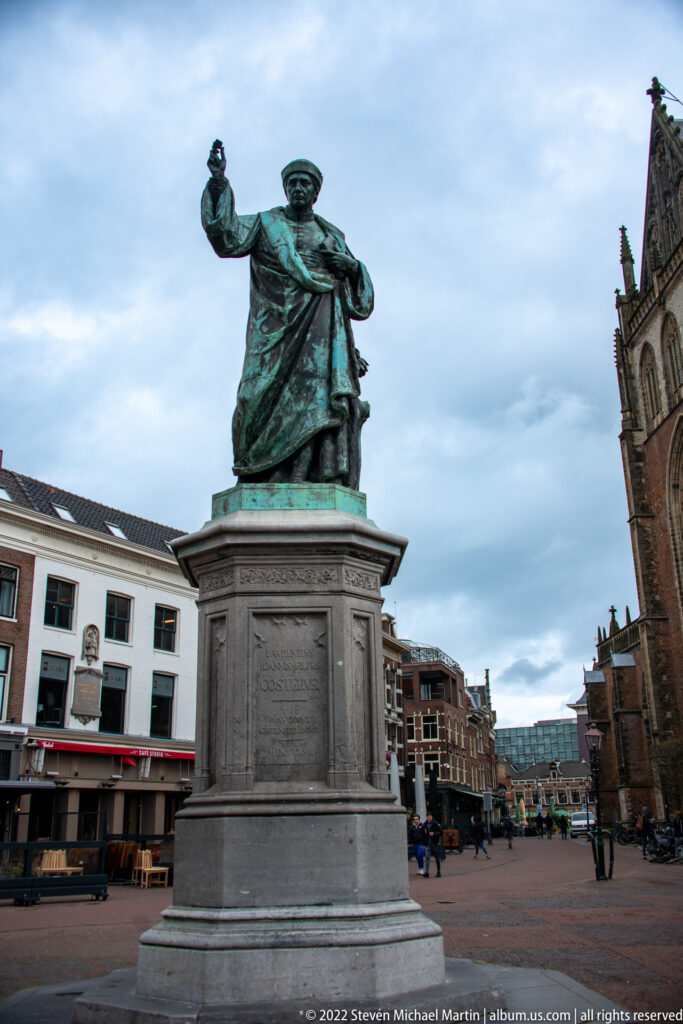 Green-patinated bronze statue of a standing man on a tall stone pedestal in a broad Haarlem square.