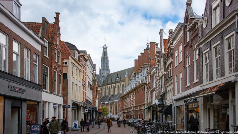 Narrow Haarlem street lined with brick shops and bicycles, leading toward the Grote or St.-Bavokerk tower in the distance.