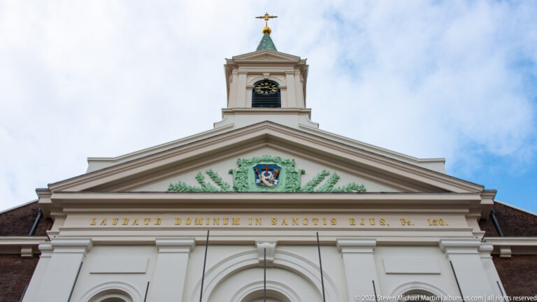 Pale Neoclassical church façade with triangular pediment, coat of arms, gilded Latin inscription, clock turret, and cross.