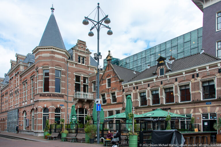 Brick Philharmonie Haarlem corner building beside historic gabled façades and a café terrace, with a modern glass structure behind.