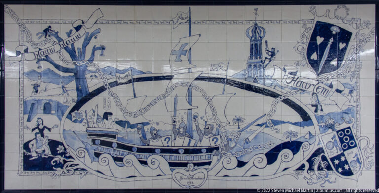 Blue-and-white tile mural with a ship, bridge, tower, heraldic shields, and decorative Haarlem imagery.