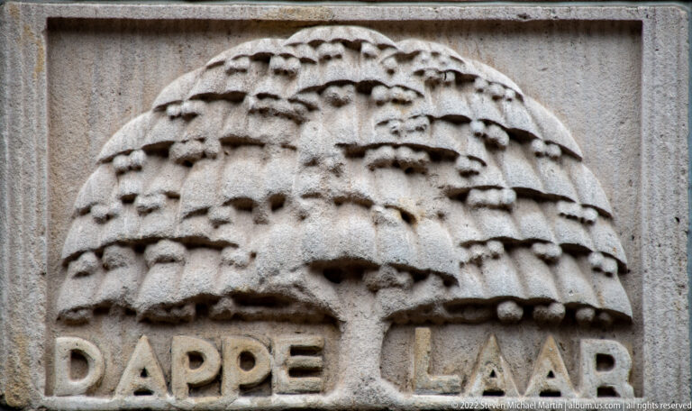Weathered stone relief of a hive-like cluster above partially visible carved letters reading “DAPPE LA…”.