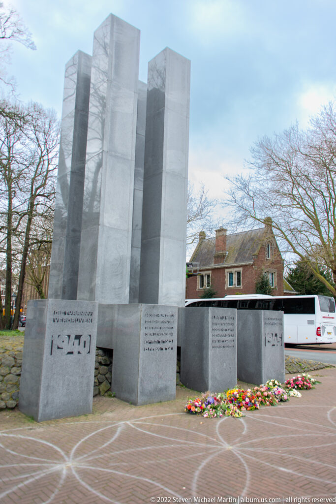 Tall pale stone memorial slabs with inscribed blocks and bouquets at the base on a paved plaza in The Hague.