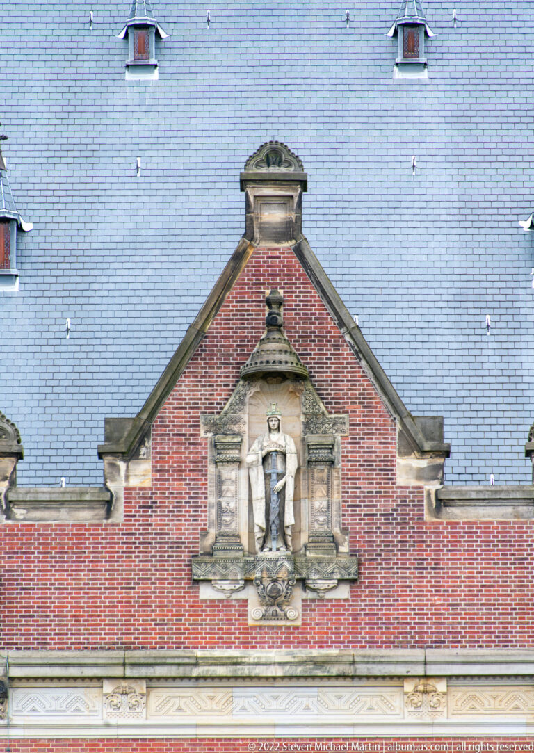 Standing stone figure in a niche within a shaped red-brick gable below the dark slate roof of the Peace Palace.