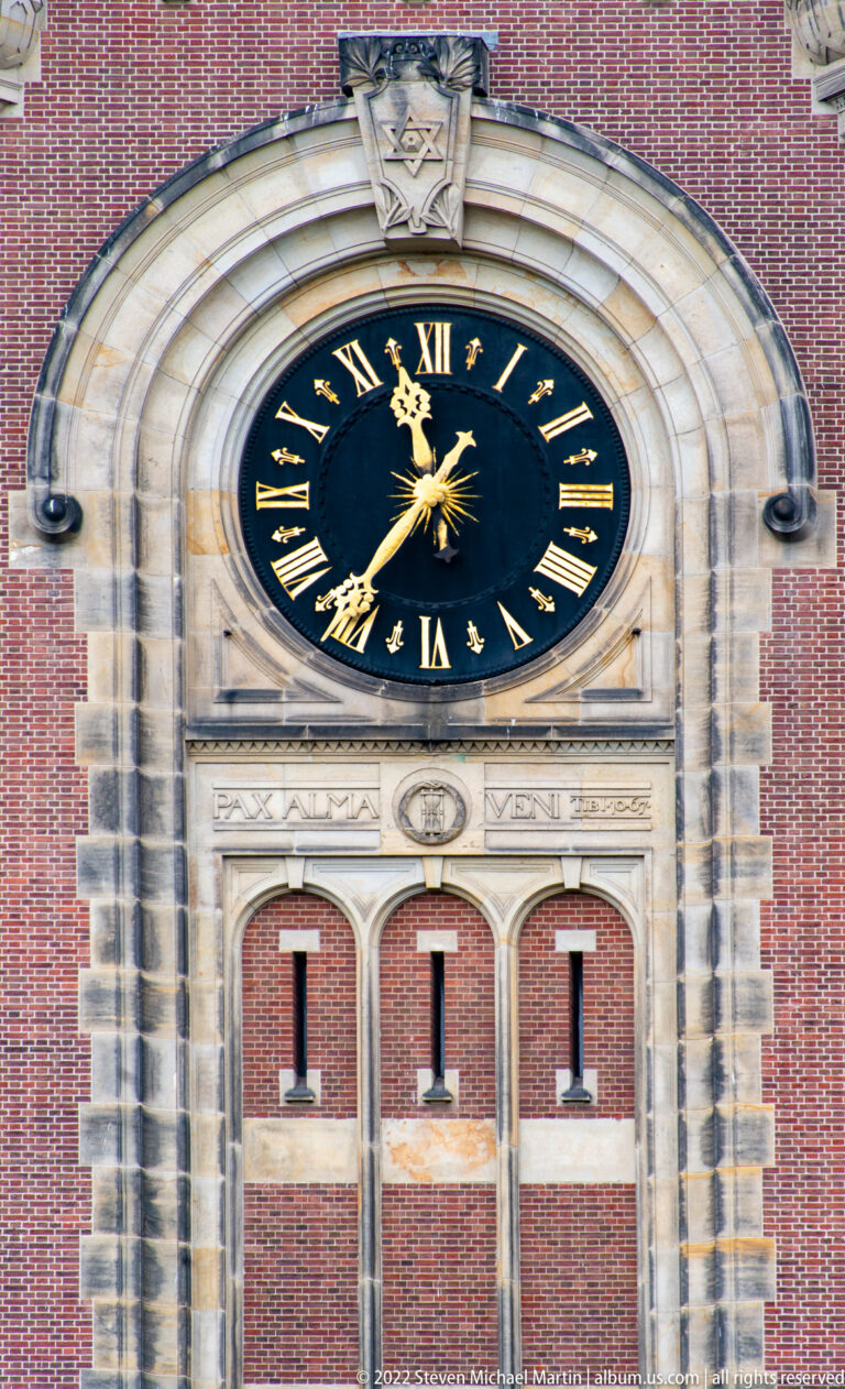Black circular clock with gold Roman numerals and hands set into pale stone on the red-brick Peace Palace tower.