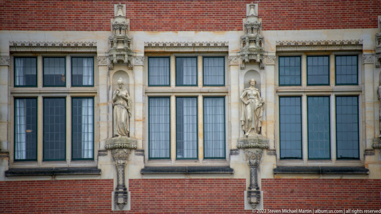 Two standing stone figures on brackets between tall windows in the red-brick Peace Palace façade.