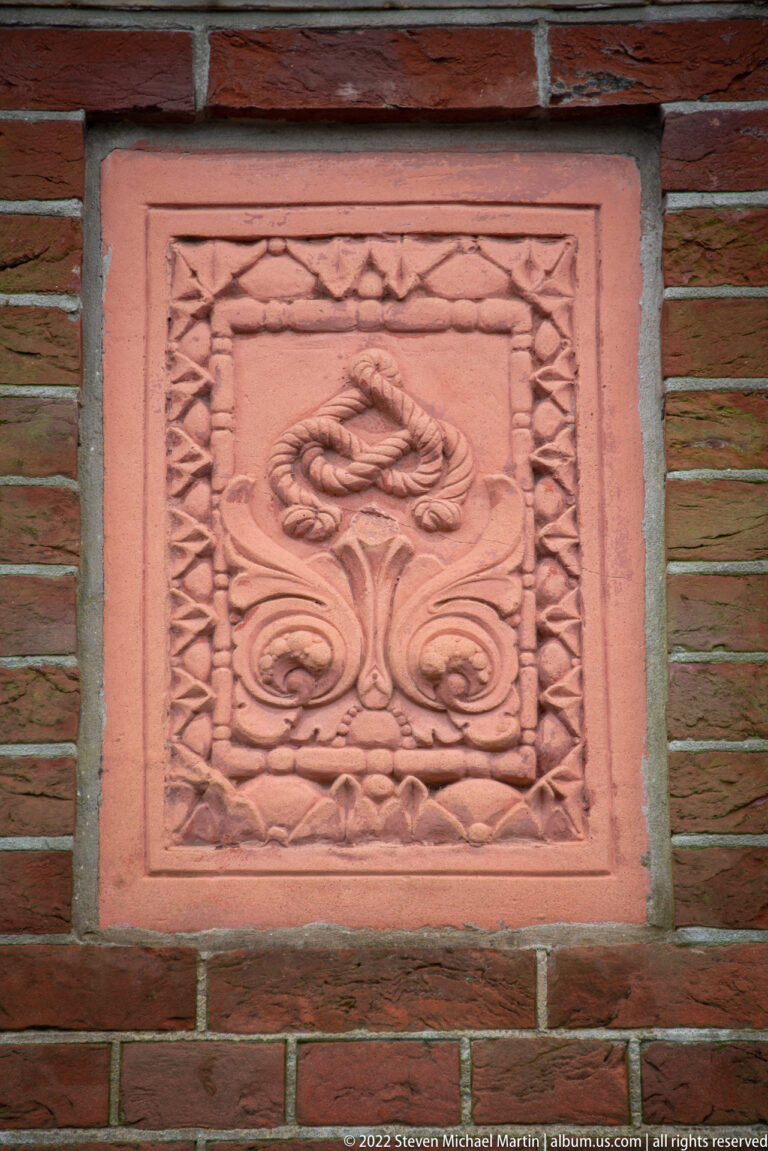 Square terracotta floral relief with curling stems embedded in a red-brick wall at the Peace Palace.