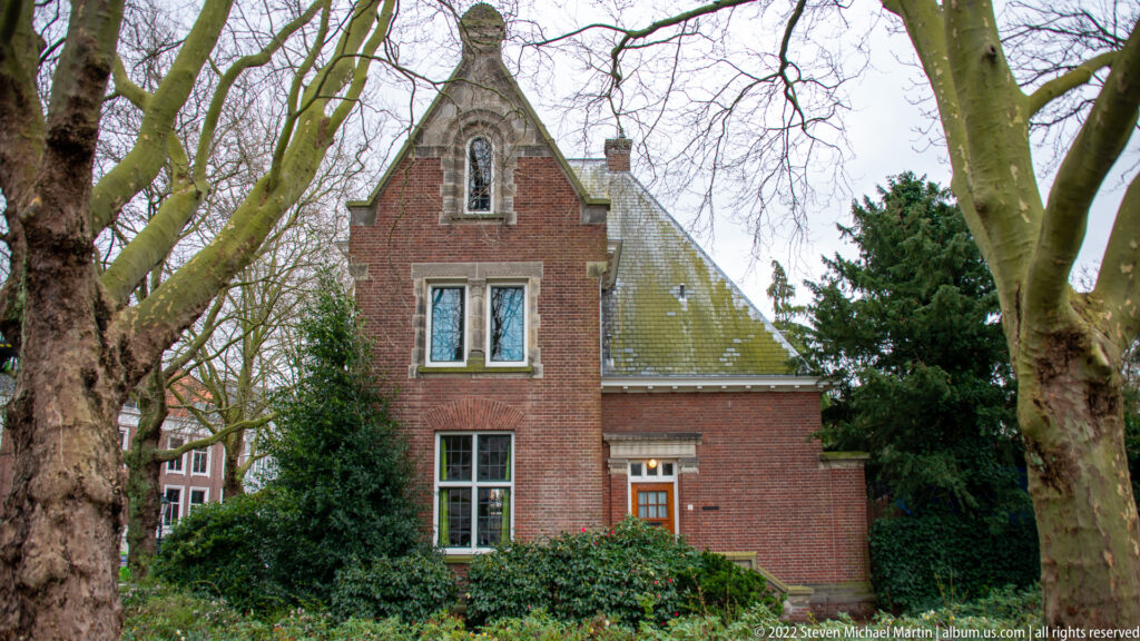 Small red-brick building with steep roof and shaped gable among mature trees and shrubs on the Peace Palace grounds.