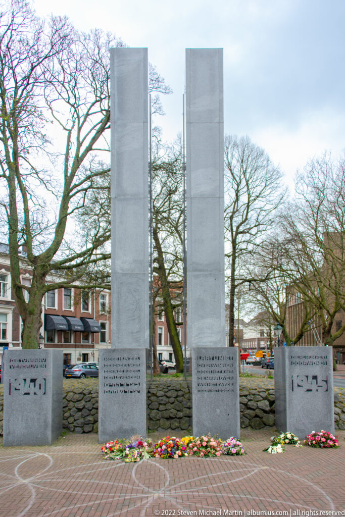 Frontal view of three tall stone memorial slabs above inscribed blocks, with flowers arranged along the base.