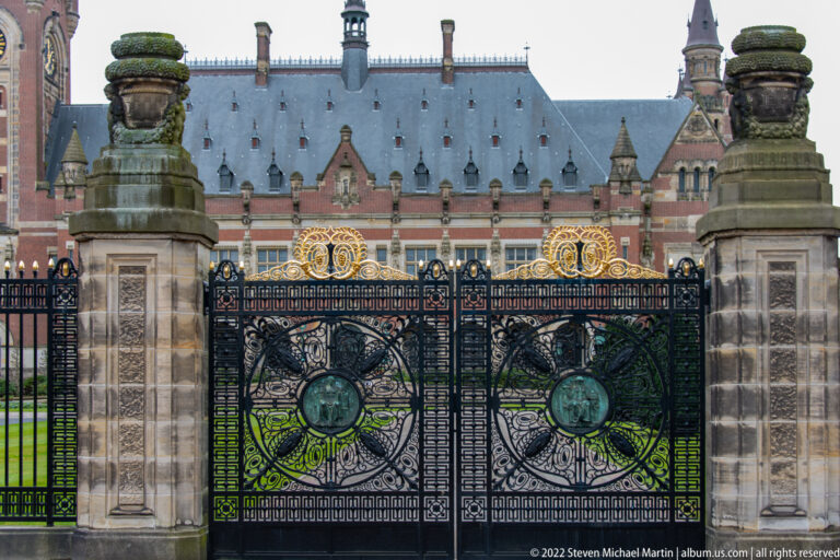 Black wrought-iron Peace Palace gate with gilded ornament, bronze medallions, stone piers, and palace façade behind.