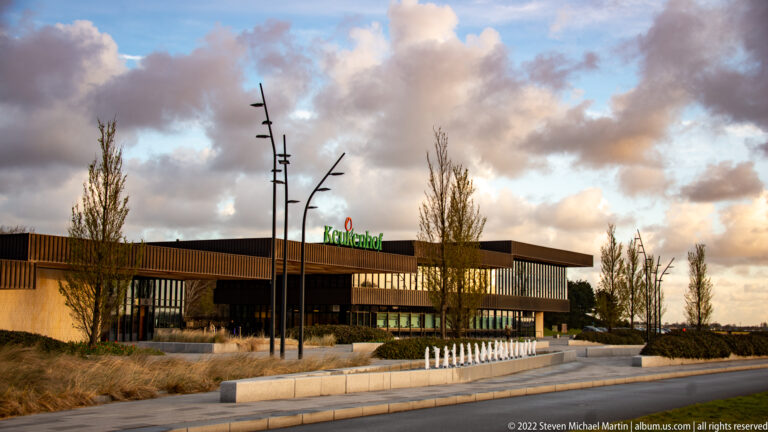 Low modern Keukenhof entrance building with paved forecourt, young trees, sculptural lights, and clouds at dusk.