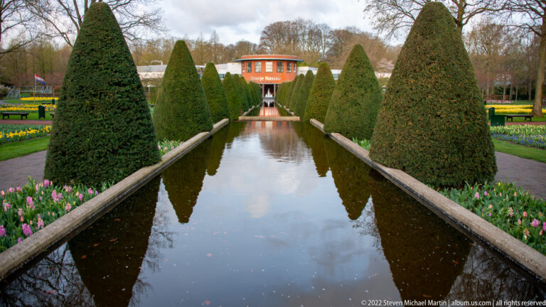 Narrow reflecting canal lined with conical topiary and spring flowers, with the Oranje Nassau pavilion at the far end.