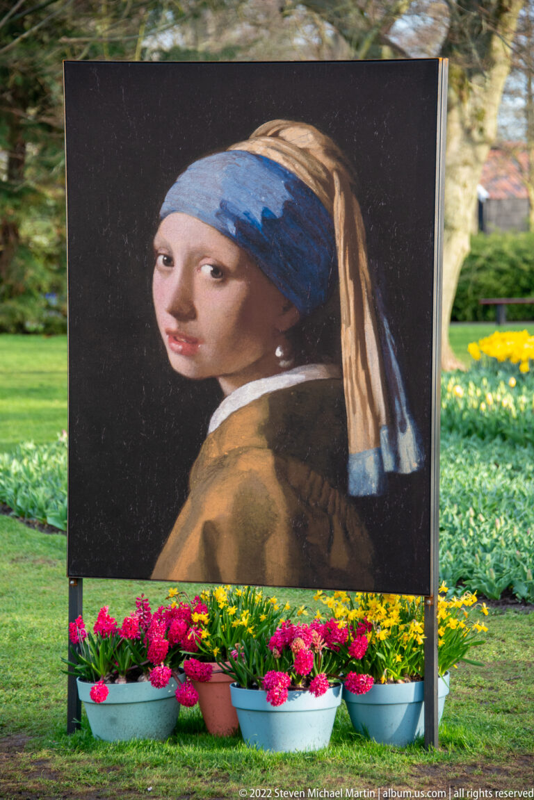 Large outdoor reproduction of Vermeer’s Girl with a Pearl Earring above pots of flowers in the garden.