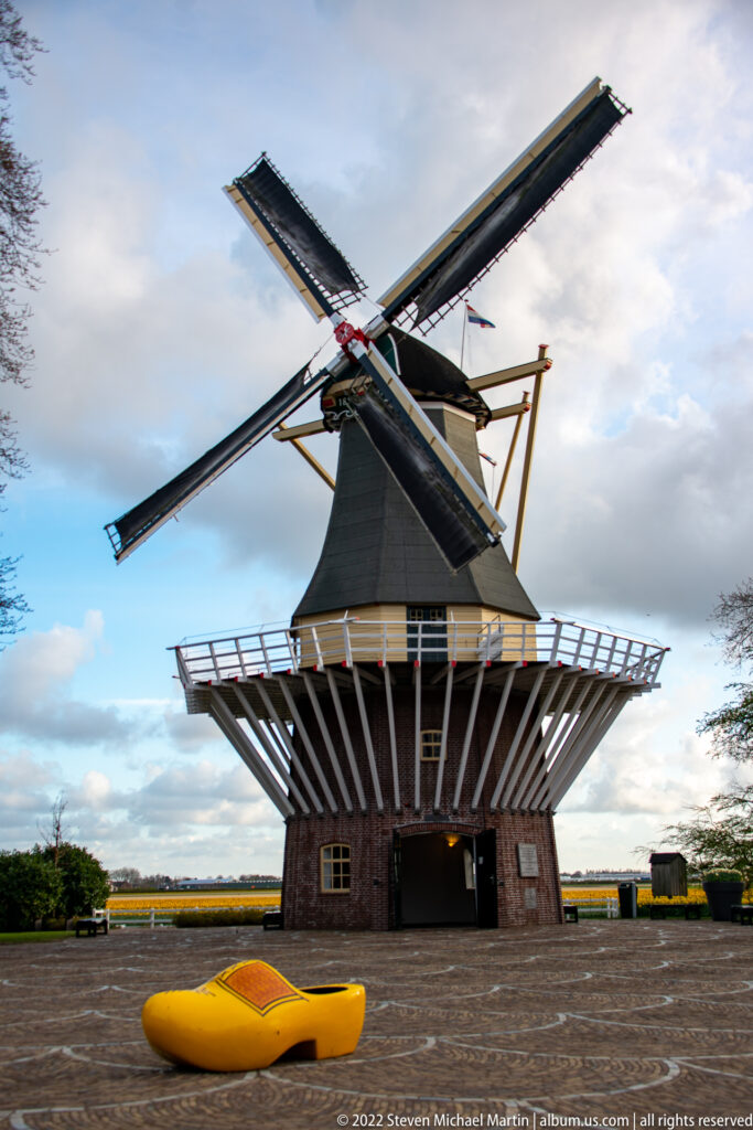 Oversized yellow wooden clog sits before the Keukenhof windmill, whose four sails fill the upper frame.
