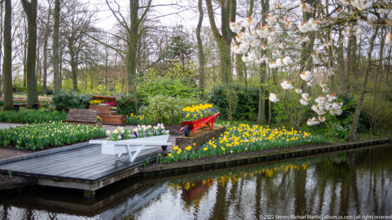 Canal beside tulip beds, white benches, colorful decorative carts, and a flowering tree in a wooded garden.