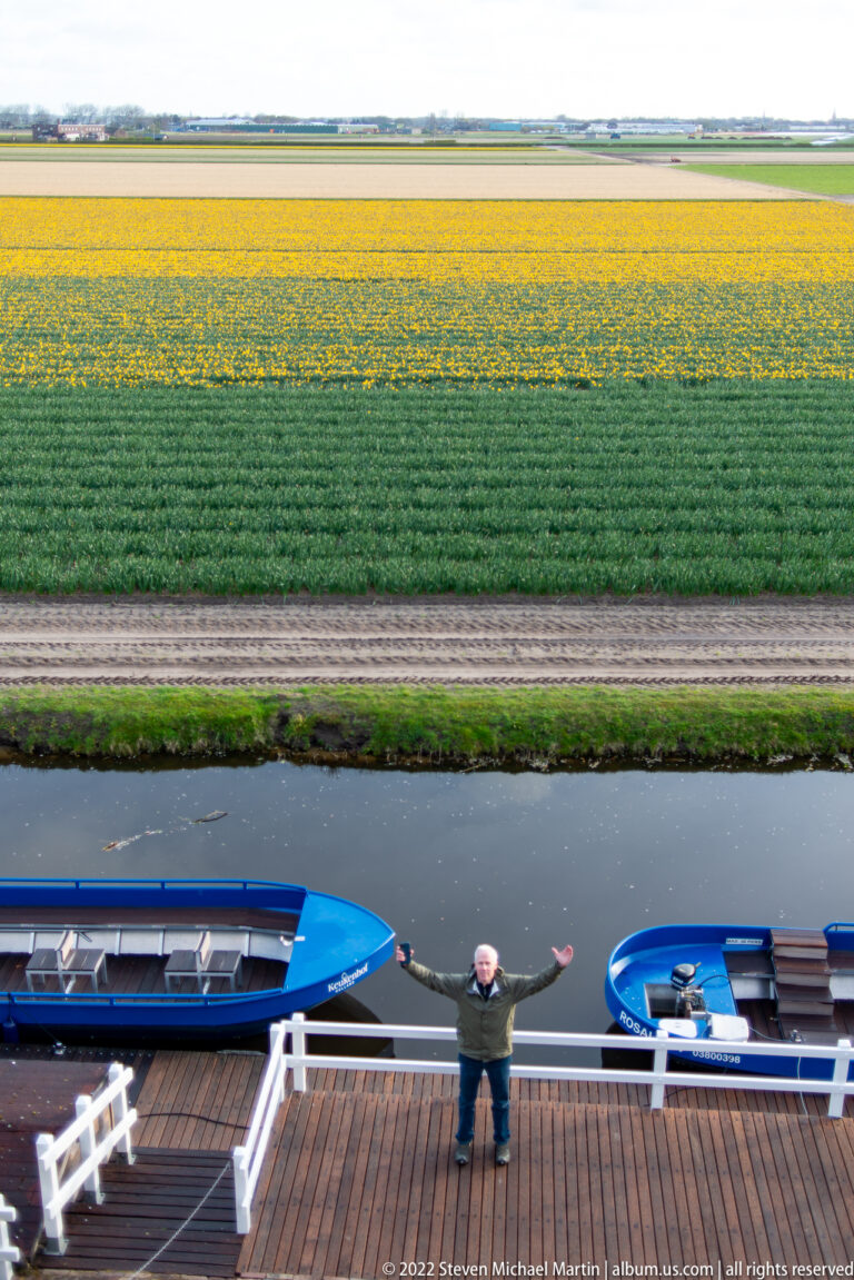 Yellow and green bulb fields beyond a canal with two blue boats and a visitor standing at a railing.
