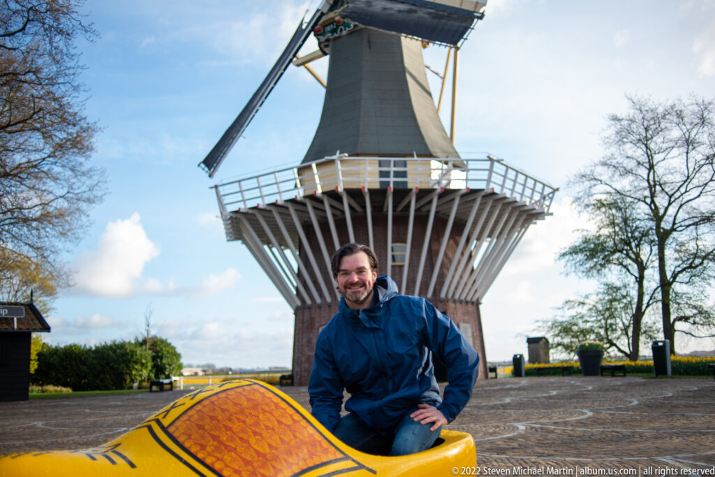 Visitor kneels beside a giant yellow clog with the Keukenhof windmill rising directly behind.