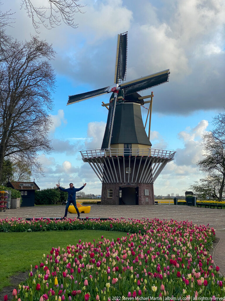 Traditional Dutch windmill behind pink and red tulip beds beneath a partly cloudy blue sky at Keukenhof.