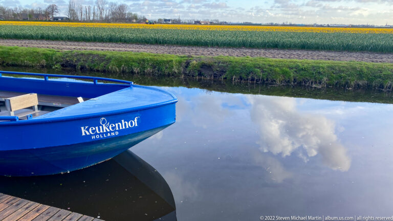 Blue boat labeled Keukenhof Holland beside a canal, with striped bulb fields extending across the opposite bank.
