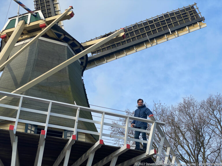 Low-angle view of a dark Dutch windmill, white gallery railing, visitor, and one sail against blue sky.
